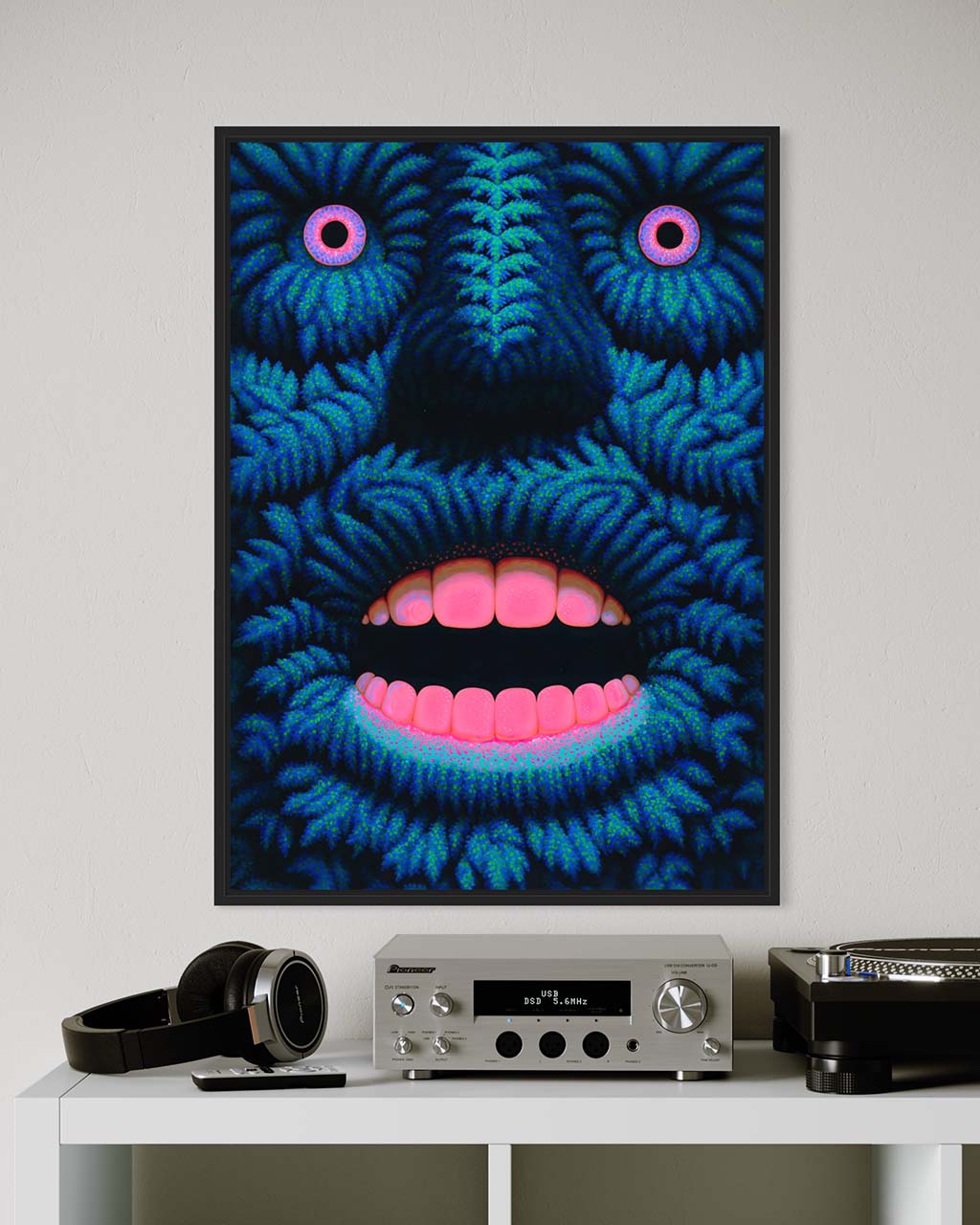 Framed artwork of a blue and pink abstract face on a white wall above a white shelf with electronic equipment.