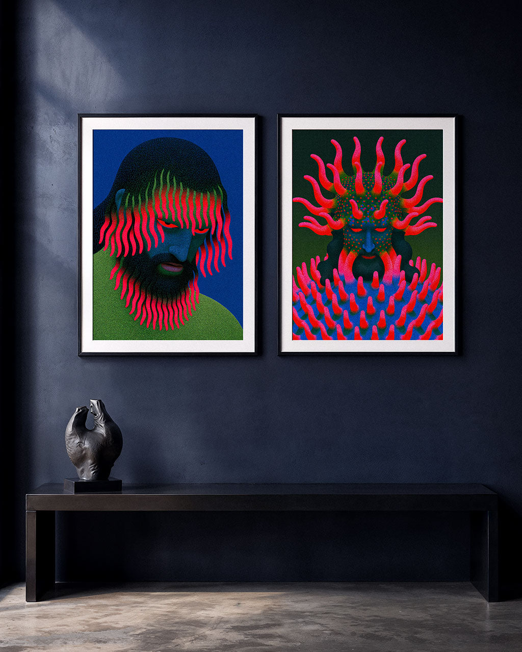 Two colourful surreal art prints displayed side by side in black frames on a dark blue wall, featuring blue-faced figures with vivid pink and green organic growths.