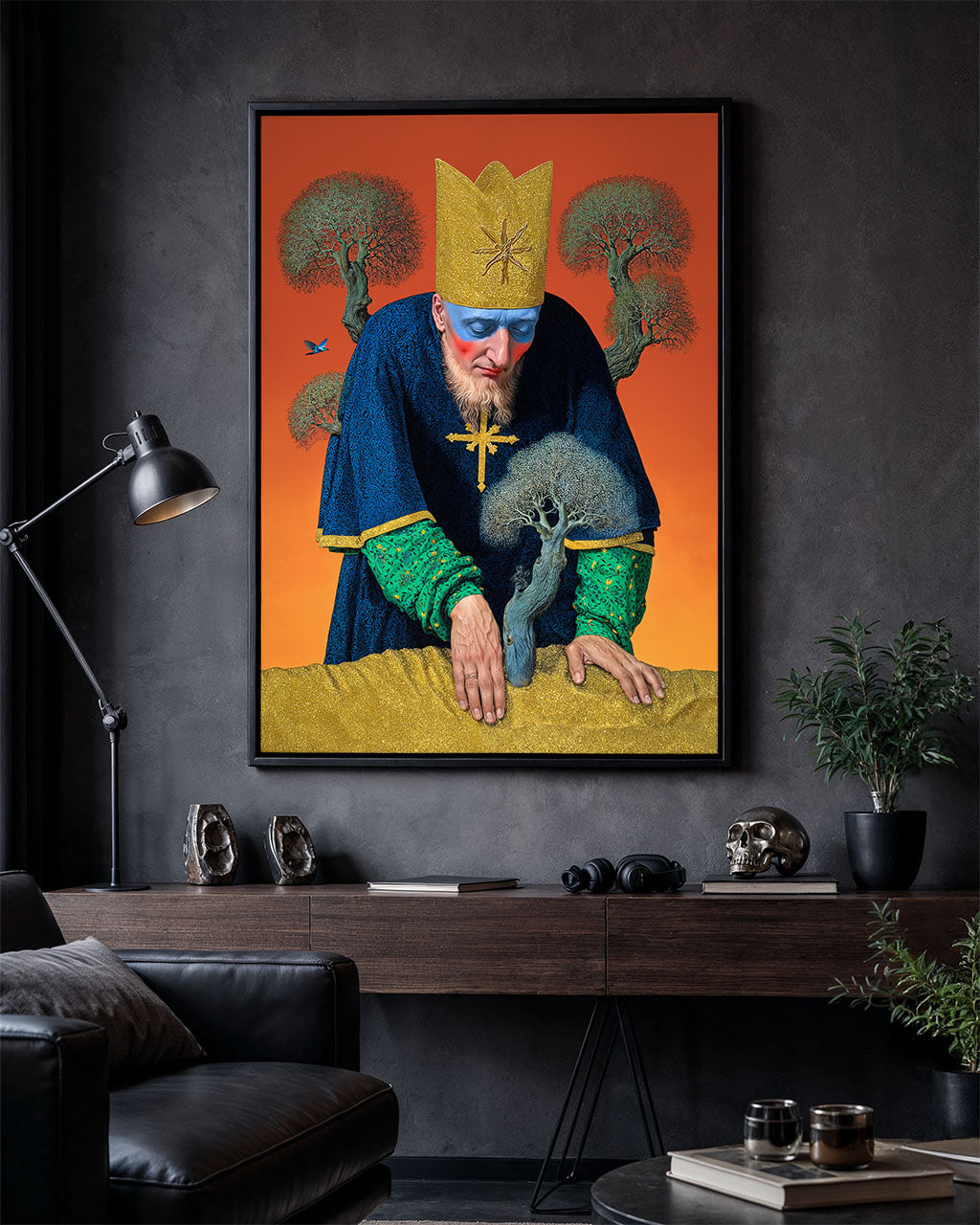Framed poster of a crowned figure leaning over a small tree, hanging on a dark wall above a wooden desk with lamp, skull and plants.
