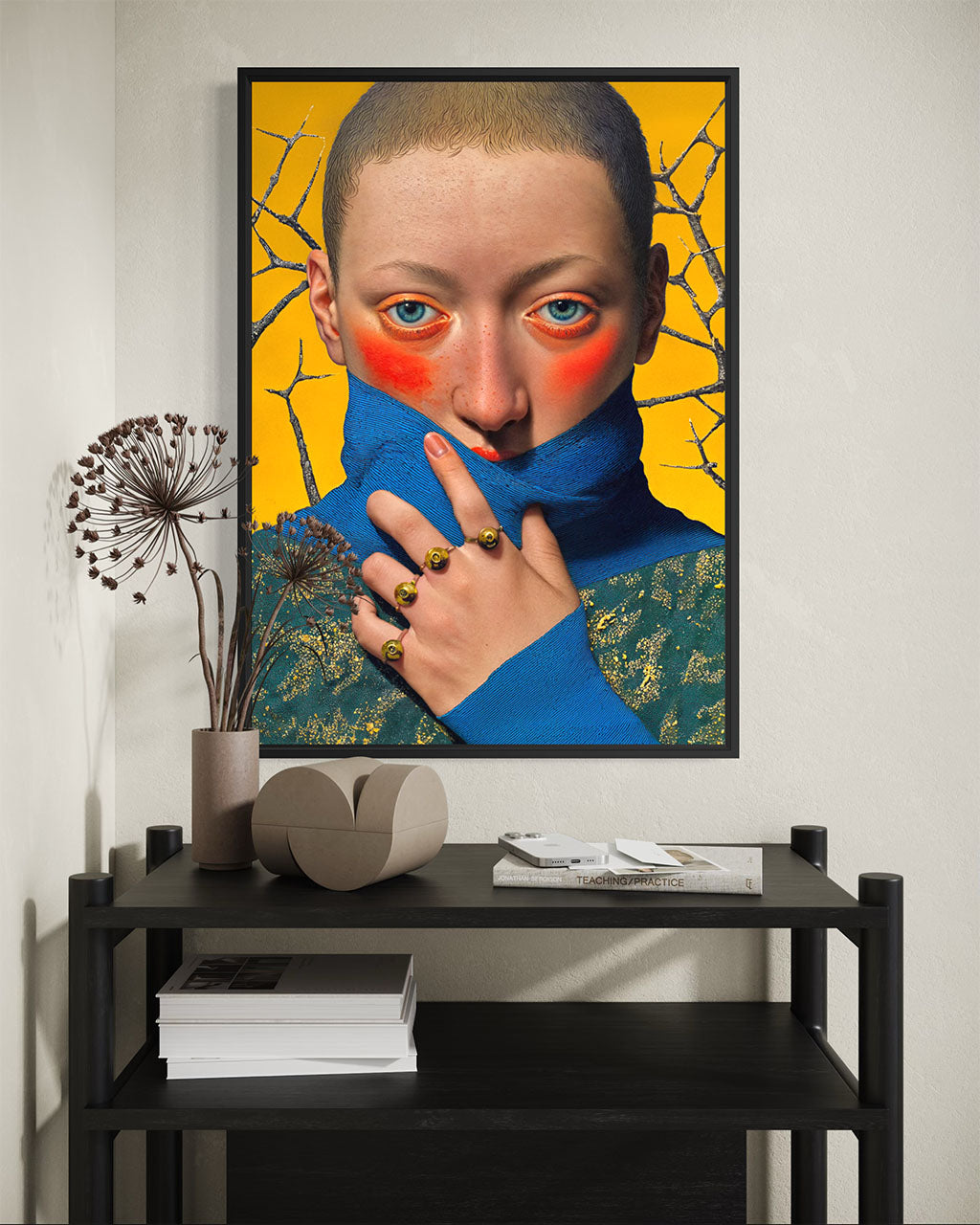 Framed poster of a blue-eyed figure with covered mouth hanging above a black shelving unit with dried flowers and stacked books.